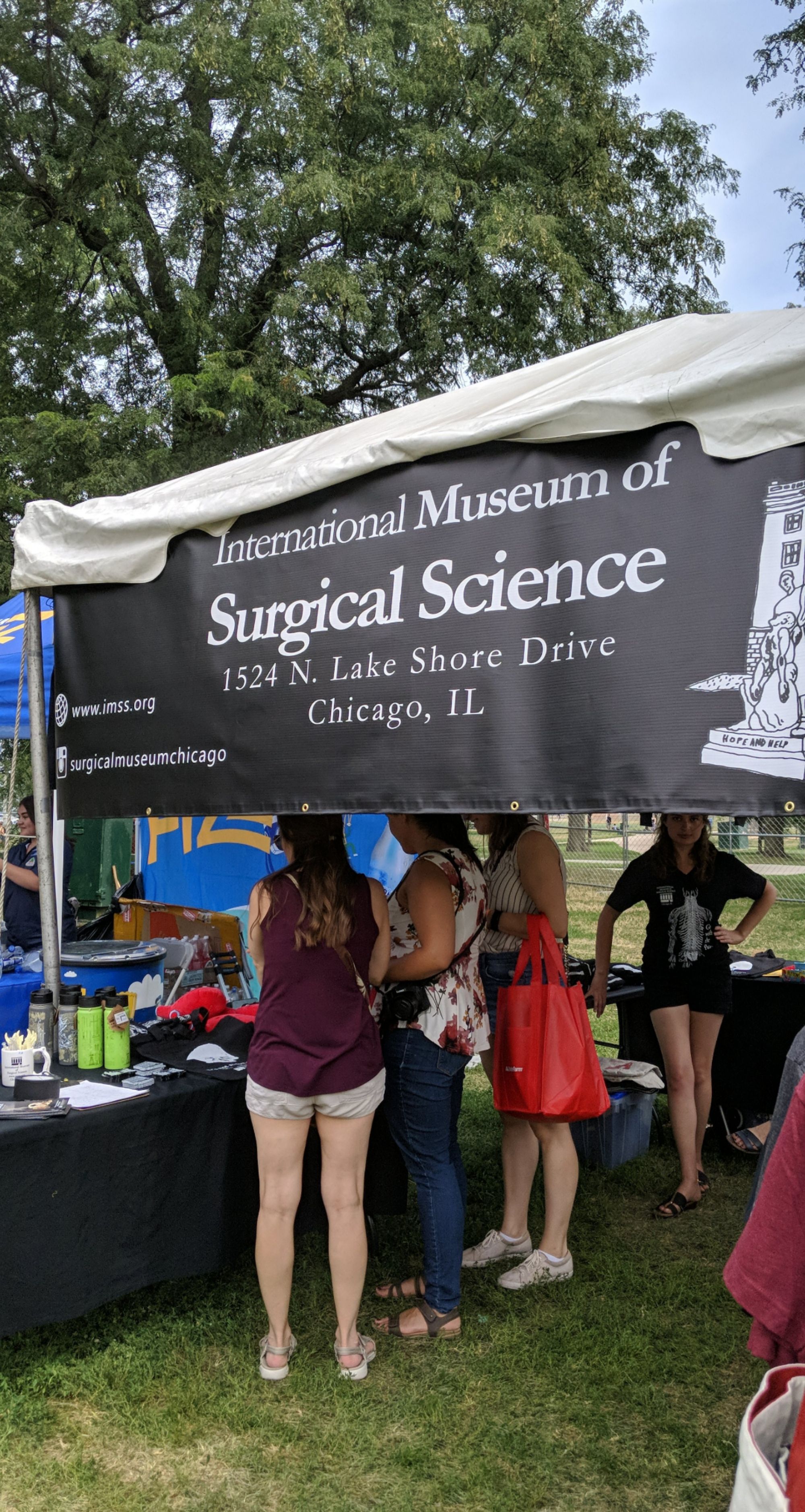 A tent with a banner reading “International Museum of Surgical Science” outdoors at the 2019 Chicago Hot Dog Fest. Visitors are looking at merchandise displayed on a table.
