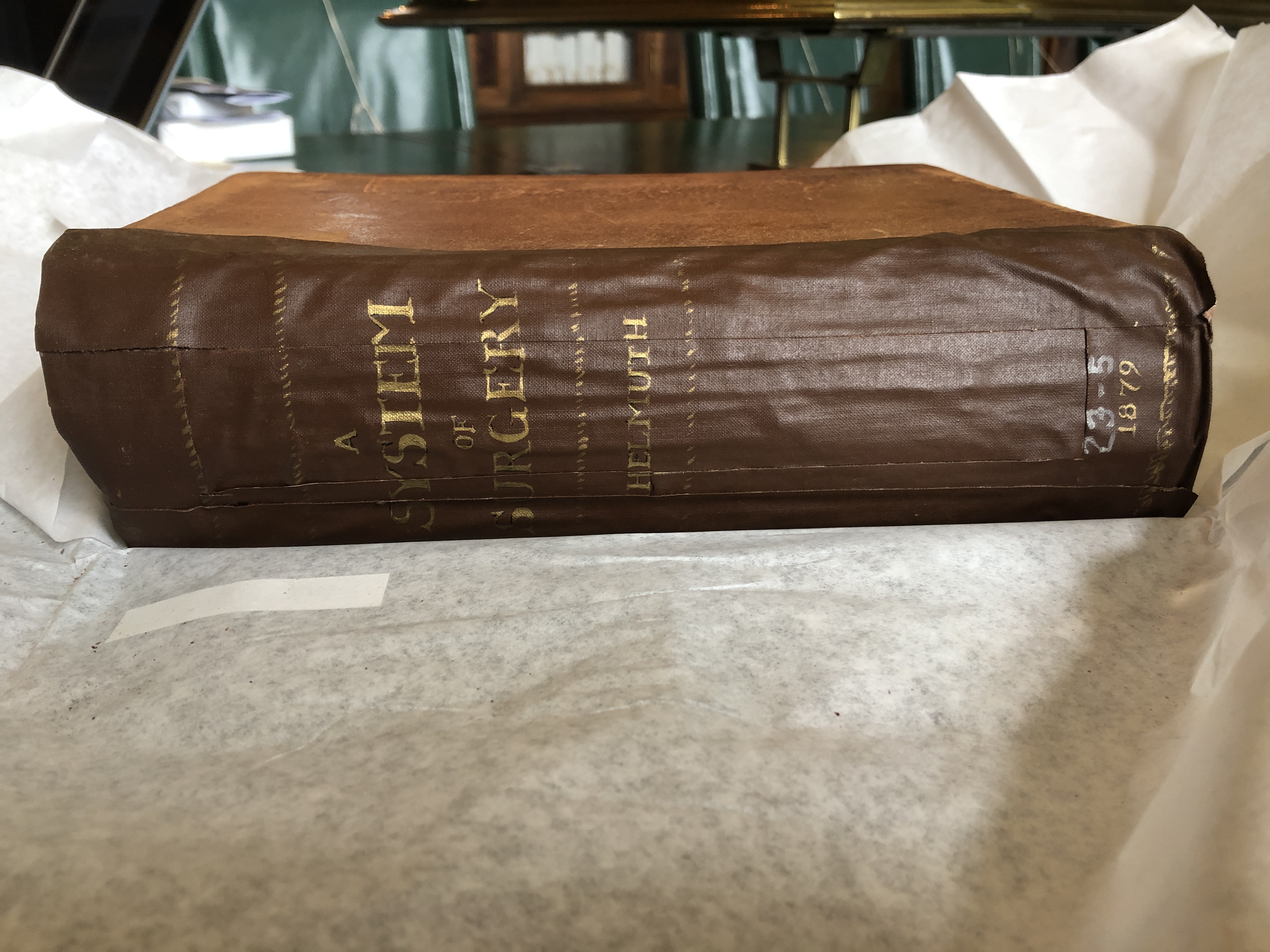 Antique brown book lays on tissue paper spread across a table with spine viewable, which reads the title of the book: “A System of Surgery” by Helmuth, and includes publication date of 1879